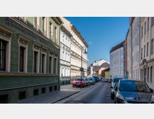 Straßenszene in einer Wohngegend mit Reihenhäusern im Altbaustil; links ein grünes Gebäude mit der Aufschrift „Frankfurter Hütte“, rechts hellere Fassaden. Am Straßenrand geparkte Autos und eine gepflasterte Gehwegfläche, bei klarem Himmel und Sonnenschein.