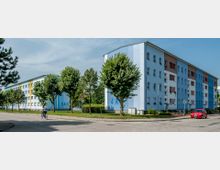 Mehrgeschossige Wohnhäuser mit hellblauer und weißer Fassade an einer Wohnstraße, umgeben von Bäumen und Grünflächen. Im Vordergrund fährt eine Person mit einem Fahrrad, daneben parkt ein rotes Auto.