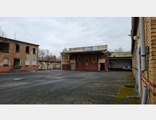 Verlassener Industriehof mit weitläufiger Asphaltfläche, umgeben von mehrstöckigen Backsteingebäuden mit beschädigten Fenstern und abgeblätterter Fassade. In der Mitte steht ein flacher Bau mit überdachtem Eingangsbereich und großen Fenstern, daneben eine offene Halle mit Graffiti.