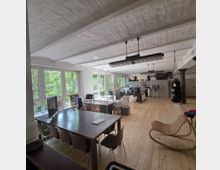 Offener Büroraum mit Holzboden, großen Fensterfronten, weißen Decken mit Backsteingewölbe und einer Mischung aus Arbeitsplätzen, Regalen und Freizeitbereichen. Im Vordergrund stehen Schreibtische mit Stühlen, im Hintergrund eine Küche, Sitzgelegenheiten und ein Boxsack.