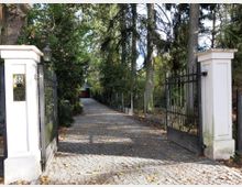 Eingangsbereich eines Grundstücks mit geöffnetem schmiedeeisernem Tor, weißen Torpfeilern und einer gepflasterten Auffahrt, die von hohen Bäumen und dichtem Grün flankiert ist. Im Hintergrund ist ein Gebäude mit Garage erkennbar.