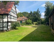 Hofplatz mit umliegenden Fachwerkhäusern, Ziegelbau und grüner Vegetation; im Vordergrund eine Wiese, rechts ein geparkter schwarzer Wagen, im Hintergrund große Bäume und ein blauer Himmel.