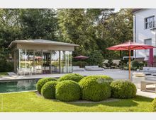 Moderner Garten mit Pool und Lounge-Bereich neben einem gläsernen Pavillon. Im Vordergrund sind gepflegte Buchsbaumsträucher, rote Sonnenschirme und Sitzmöbel auf einer Terrasse sichtbar, umgeben von dichtem Baumbestand.