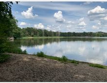Sandstrand an einem ruhigen See, der von Bäumen und dichter Vegetation umringt ist. Im Hintergrund spiegeln sich Wolken im Wasser unter klarem, teils bewölktem Himmel.