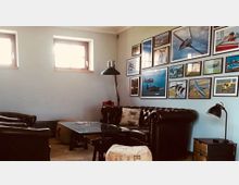 Ein gemütlicher Raum mit Ledersofas, einem Glastisch und zwei Fenstern mit Holzrahmen. An einer Wand hängt eine Sammlung von gerahmten Bildern und Postern, die Flugzeuge und Luftfahrtmotive zeigen.