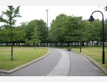 Ein Park mit einer gepflasterten Gehwegkurve, umgeben von Rasenflächen und zahlreichen Bäumen. Schwarze Laternen und grüne Sitzbänke säumen den Gehweg, im Hintergrund sind weitere Bäume und eine städtische Umgebung erkennbar.