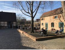 Ein gepflasterter Innenhof mit Backsteingebäuden, darunter ein Haus mit blauen Fensterläden und ein Fachwerkhaus. Vor einem Baum sitzen Menschen an Tischen auf einer erhöhten Holzterrasse, umgeben von ländlicher Atmosphäre und kahlen Bäumen.