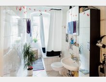 Bright, narrow tiled bathroom with a bathtub and white shower curtain at the far end beneath a large window. A wall-mounted sink with mirrored cabinet, towels, a striped floor rug, and potted plants line the space.