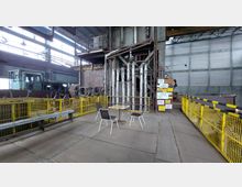 Industrielle Halle mit Metallflächen und Rohren, gelben Sicherheitsgittern und Beschilderungen. Im Vordergrund steht eine kleine Sitzgruppe mit einem Tisch und zwei Stühlen.