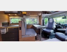 Innenraum eines modernen Wohnmobils mit Sitzbereich, Klapptisch und drehbarem Fahrersitz. Links eine kompakte Küchenzeile mit Holzverkleidung, darüber Schränke und ein Fenster, das ins Grüne blickt.