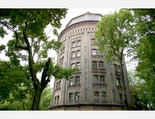 Rundes, mehrstöckiges Gebäude aus hellem Backstein mit zahlreichen Fenstern und verzierten Fassadendetails, umgeben von hohen Bäumen in einer parkähnlichen Umgebung.