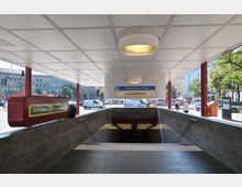 Eingang der U-Bahn-Station "Fehrbelliner Platz" in einer urbanen Umgebung, mit überdachtem Bereich, einem blau-weißen Stationsschild und einer Treppe, die nach unten führt. Im Hintergrund sind Gebäude, Bäume und vorbeifahrende Autos sichtbar.