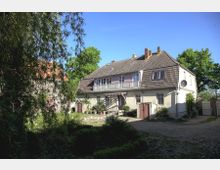 Ein zweigeschossiges Wohnhaus mit heller Fassade und schrägem Dach, umgeben von Pflanzen, Bäumen und einem gepflasterten Hof. Im Hintergrund ist ein weiteres Gebäude mit rotem Ziegeldach sichtbar.