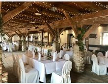 Rustikaler Veranstaltungsraum mit Holzbalkendecke, freiliegenden Ziegelmauern und dekorativen Lichterketten. Die Tische sind mit weißen Tischdecken, Stuhlhussen und schlichten Blumengestecken für eine Feier eingedeckt.