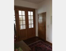 Ein kleiner Eingangsbereich mit Holztüren; eine doppelflügelige Glastür mit Holzrahmen auf der linken Seite und eine schlichte Holztür rechts daneben. Der Boden ist aus dunklem Holz, teils bedeckt von einem farbigen Teppich.