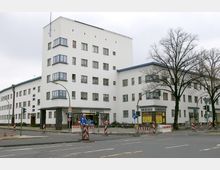 Mehrstöckiges Wohn- und Geschäftsgebäude im Stil der Moderne mit weißer Fassade und zahlreichen rechteckigen Fenstern. Im Erdgeschoss befinden sich Geschäfte, Baustellenabsperrungen und Straßenschilder prägen den Bereich vor dem Gebäude.