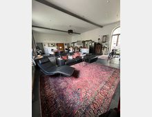 Großzügiger Wohnraum mit offener Küche, schwarzen Ledermöbeln, einem großen roten Orientteppich und einer Liege. Im Hintergrund stehen Küchenmöbel, ein großer Kühlschrank und eine hohe Fensterfront mit halbkreisförmigem Oberlicht.