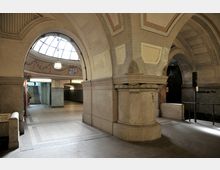 Historischer U-Bahnhof mit hohen Gewölbedecken, verzierten Bögen und einem zentralen Lichteinlass im Kuppelstil. Im Hintergrund führen Treppen zu einem Ausgang, und ein Schild zeigt den Stationsnamen "Heidelberger Platz".
