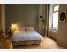Schlafzimmer mit einem großen Bett in der Mitte, weißer Bettwäsche und beigen Kissen. Links steht eine Nachttischlampe auf einem Tisch mit einer Topfpflanze, rechts hängt eine Zimmerpflanze in einem Makramee, und ein Fenster mit weißen Vorhängen lässt Tageslicht herein.