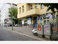 Straßenszene mit einer gelben Gebäudefassade, die bunte Wandmalereien mit abstrakten und futuristischen Motiven zeigt. Im Vordergrund stehen ein Stromkasten mit Plakaten, ein geparkter weißer Motorroller und Fahrräder; Straßenschilder weisen auf die Kreuzung Dunckerstraße hin.