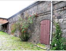 Steingebäude mit grober Steinmauer und roten Holztüren, flankiert von Kletterpflanzen mit roten Blüten. Im Vordergrund ein gepflasterter Hof mit etwas Grasbewuchs.