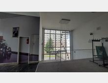 Innenraum eines modernen Gebäudes mit einer großen Fensterfront, die den Blick auf einen Hinterhof und ein Wohngebäude bietet. Im Vordergrund befinden sich ein Treppengeländer, eine Garderobe, eine Tür mit Feuerlöscher und eine mit Pflanzen dekorierte Wand.