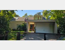 Modernes Einfamilienhaus mit Flachdach und hell beiger Fassade, einer Garage, einem großen Fenster im Obergeschoss und einem Basketballkorb im Vorgarten. Das Haus ist umgeben von dichtem Grün und einem schwarzen Metallzaun im Vordergrund.