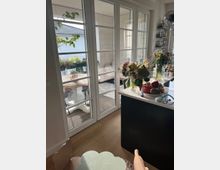 Ein Wohnraum mit Holzboden und großen Glas-Doppeltüren, die zu einer Terrasse mit Holzboden und Tisch führen. Im Innenraum stehen Blumen auf einer Kücheninsel, dahinter befinden sich Regale mit Gläsern und Lebensmitteln.