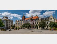Historischer Marktplatz mit Pflastersteinen, umgeben von Gebäuden verschiedener Architekturstile, darunter ein großes Gebäude mit rotem Satteldach und Turm mit Uhr. In der Platzmitte steht ein geschmückter Maibaum, umgeben von Bäumen, Blumenkübeln und einigen Passanten.