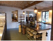 Industrieküche in einem Loft mit freiliegenden Backsteinwänden und sichtbaren Holzbalken. Im Vordergrund eine große Arbeitsplatte mit integriertem Kochfeld, darunter Geschirrregale; im Hintergrund ein Fenster mit schwarzem Metallrahmen und Blick ins Grüne.