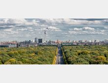 Panoramablick auf Berlin mit dem Fernsehturm im Zentrum, der von modernen und historischen Gebäuden umgeben ist, im Vordergrund ein von Bäumen gesäumter Straßenzug durch den Tiergarten.