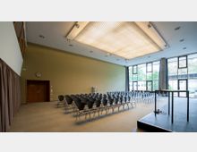 Moderner Konferenzraum mit Reihen schwarzer Stühle, einem erhöhten Podium mit Mikrofon und großer Fensterfront, die Tageslicht hereinlässt. An der linken Seite befindet sich eine Holztür mit der Aufschrift „K1“ und eine Wanduhr.