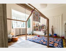 Ein helles Kinderzimmer mit einem großen, hölzernen Spielhausrahmen in der Mitte, an dem gemalte Kinderbilder befestigt sind. Im Hintergrund befinden sich ein Bücherregal, ein bunt gemusterter Teppich und ein Erkerfenster mit weißen Sprossen und grünen Vorhängen.