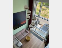 Wohnbereich mit hohen Decken, großen bodentiefen Fenstern und Blick auf eine Straße mit Grünfläche. Im Raum befinden sich eine rosa Couch, ein goldener runder Hocker, Hängelampen mit kugelförmigen Leuchten, ein modernes Gemälde und Zimmerpflanzen.