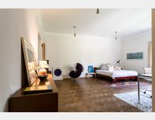 Schlafzimmer mit Holzboden, weiß gestrichenen Wänden und minimaler Einrichtung. Im Raum stehen ein holzgerahmtes Bett mit gemusterten Kissen auf einem bunten Teppich, ein lila Sessel, ein niedriger Holztisch sowie ein Sideboard mit dekorativen Objekten und Kunstwerken.