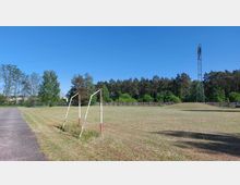 Eine einfache Wiese mit einem verrosteten Fußballtor im Vordergrund, umgeben von einer Baumreihe und einem Zaun. Im Hintergrund ist ein Wald sowie ein hoher Strommast zu sehen, der sich vor einem klaren, blauen Himmel erstreckt.