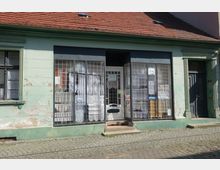 Fassade eines kleinen, alten Gebäudes mit grün gestrichenen Wänden und abgeblätterter Farbe. Im Erdgeschoss befinden sich große Schaufenster mit Metallgittern, hinter denen Spitzenvorhänge und Kartons sichtbar sind; daneben eine Tür mit Glasscheibe.