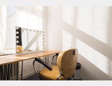 Ein minimalistischer Schminktisch mit einem großen beleuchteten Spiegel, der von runden Glühbirnen umrahmt ist, steht in einem hellen Raum mit weißen Wänden und Tageslicht. Ein gepolsterter Lederstuhl im Vintage-Stil steht davor.