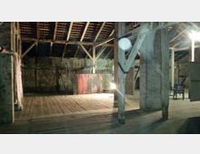 Ein leerer Dachbodenraum mit sichtbaren Holzbalken und altem Holzfußboden. Im Hintergrund steht eine Staffelei mit einem Gemälde, und eine schwache Beleuchtung erhellt den Raum teilweise.