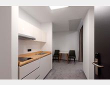 Kleine Küchenzeile mit weißen Schränken, Holz-Arbeitsplatte, Spüle und Kochfeld; im Hintergrund ein kleiner Esstisch mit zwei Stühlen auf einem grauen Teppichboden. Die Szene befindet sich in einem schlichten, hellen Raum mit glatten Wänden und dunklem Vorhang.