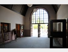 Großer Eingangsbereich mit einer hohen, gewölbten Decke und einem großen Bogenfenster mit Glaselementen, durch das Tageslicht einfällt. Offene Holztüren führen nach draußen, im Inneren sind dekorative Elemente und Fenster zu sehen.