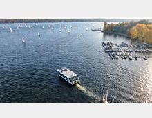 Luftaufnahme eines Sees mit vielen kleinen Segelbooten, einem modernen Motorboot im Vordergrund und einem Yachthafen mit angedockten Booten am rechten Ufer. Am Ufer stehen herbstlich gefärbte Bäume, die eine parkähnliche Landschaft umrahmen.