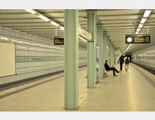 U-Bahn-Station Strausberger Platz mit einer hell beleuchteten Halle, grünen Säulen, gefliesten Wänden und Stahlbänken. Ein Zug ist nicht sichtbar; einige Menschen befinden sich auf dem Bahnsteig, und Hinweistafeln mit Abfahrtszeiten und einer Uhr sind erkennbar.
