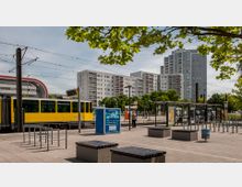 Straßenbahnhaltestelle mit gelber Straßenbahn im Vordergrund, umgeben von Sitzbänken und überdachten Wartebereichen. Im Hintergrund stehen mehrstöckige Wohngebäude mit Balkonen, umgeben von Bäumen.