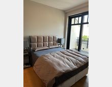 Schlafzimmer mit Doppelbett und gepolstertem Kopfteil, flankiert von zwei Nachttischen mit quadratischen Lampen. Rechts ein großes, schwarz gerahmtes Fenster bzw. eine Balkontür mit Blick auf einen kleinen Balkon mit Metallgeländer.