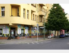 Gelbes Wohngebäude mit mehreren Balkonen und weißen Fenstern an einer Straßenecke. Der Erdgeschossbereich ist mit bunten Wandmalereien und Schildern gestaltet, daneben stehen eine Straßenlaterne, ein Baum sowie parkende Autos und Fahrräder.