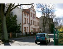 Straßenansicht eines historischen Gebäudes mit heller Fassade und roten Backsteinakzenten, das von Bäumen umgeben ist. Vor dem Gebäude steht ein geparktes blaues Auto, und im Vordergrund ist ein Metallzaun zu sehen.