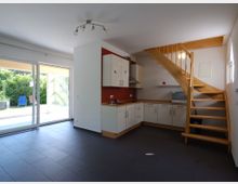 Offener Raum mit moderner Einbauküche und Holztreppe, die zu einem Obergeschoss führt. Große Glasschiebetüren bieten Zugang zu einer Terrasse und einem Garten mit grüner Hecke im Hintergrund.