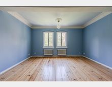 Leerer Wohnraum mit hellblauen Wänden, breiten Holz-Dielenboden und weißer Stuckleiste an der Decke. An der Stirnwand befinden sich zwei hohe Fenster über zwei weißen Heizkörpern, mittig hängt eine runde Deckenleuchte.
