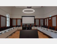 Konferenzraum mit oval angeordneten Tischen, Mikrofonen auf den Arbeitsplätzen und schwarzen Stühlen. Im Hintergrund befindet sich eine große Leinwand, umgeben von holzgerahmten Akustikpaneelen; die Decke ist hoch mit einem modernen, kreisförmigen Leuchtring als Beleuchtung.